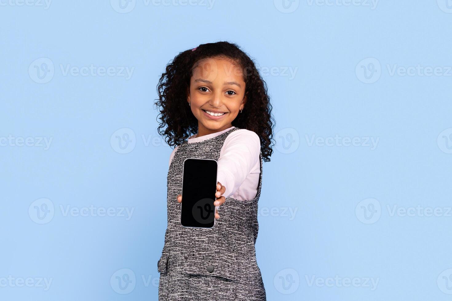 A cheerful girl with curly hair holds out a smartphone, smiling widely. She is dressed in a stylish grey outfit, surrounded by a bright blue backdrop. photo