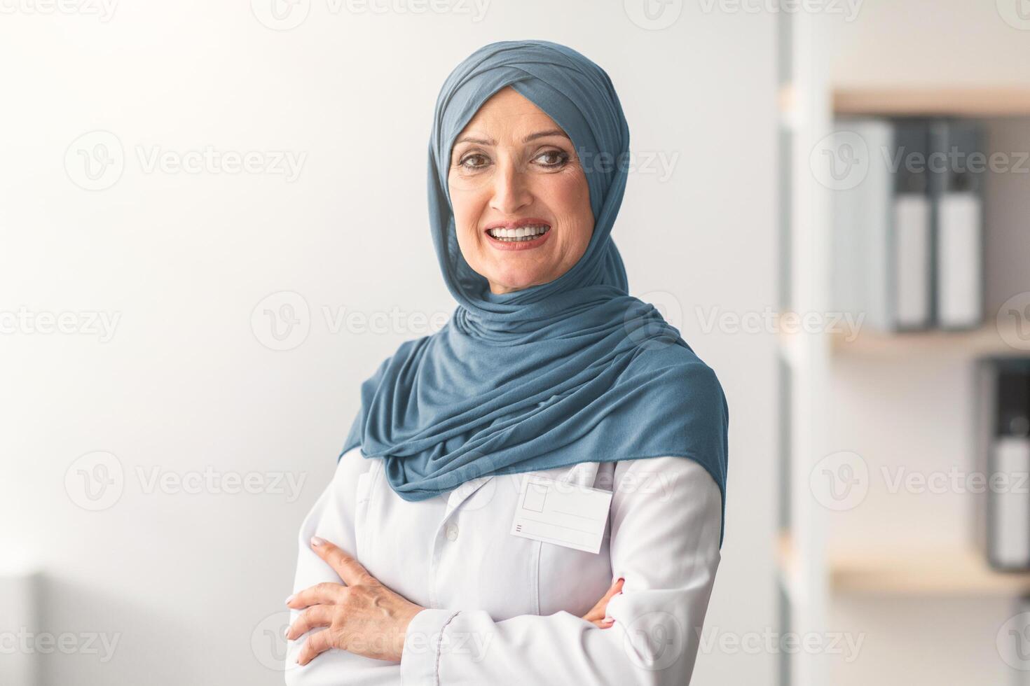 Mature Muslim Doctor Woman Wearing Hijab Smiling To Camera Standing Crossing Hands In Clinic Office. Portrait Of Professional Therapist Arab Lady Concept. Free Space For Text photo