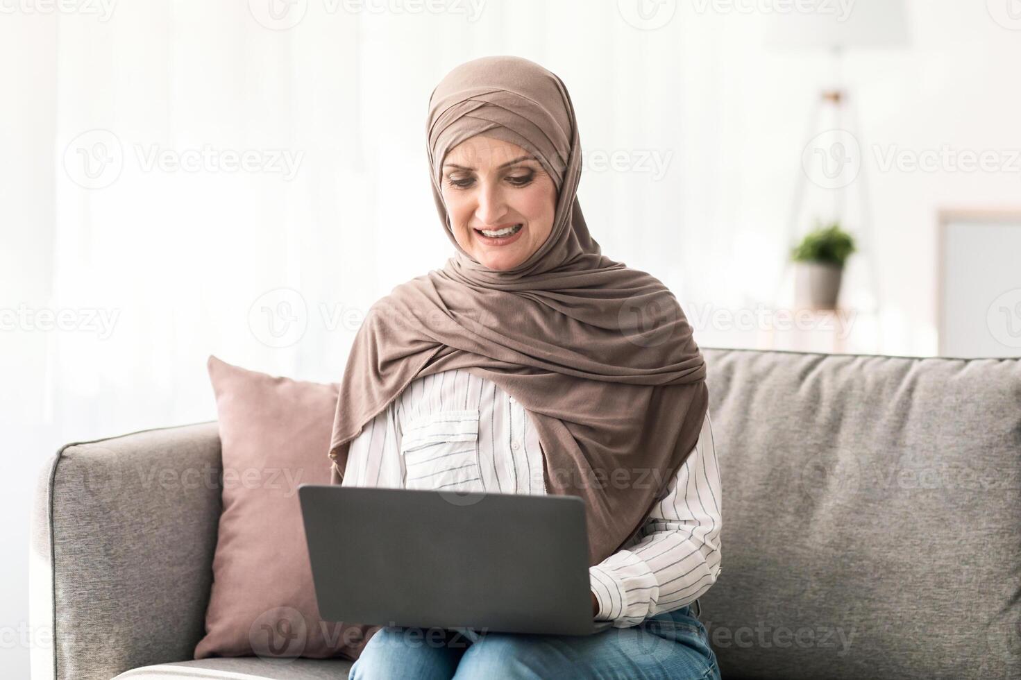 Mature Middle-Eastern Woman In Hijab Using Laptop Computer Browsing Internet Sitting On Couch At Home. Modern Muslim Lady Working Distantly Online Indoors. People And Gadgets photo