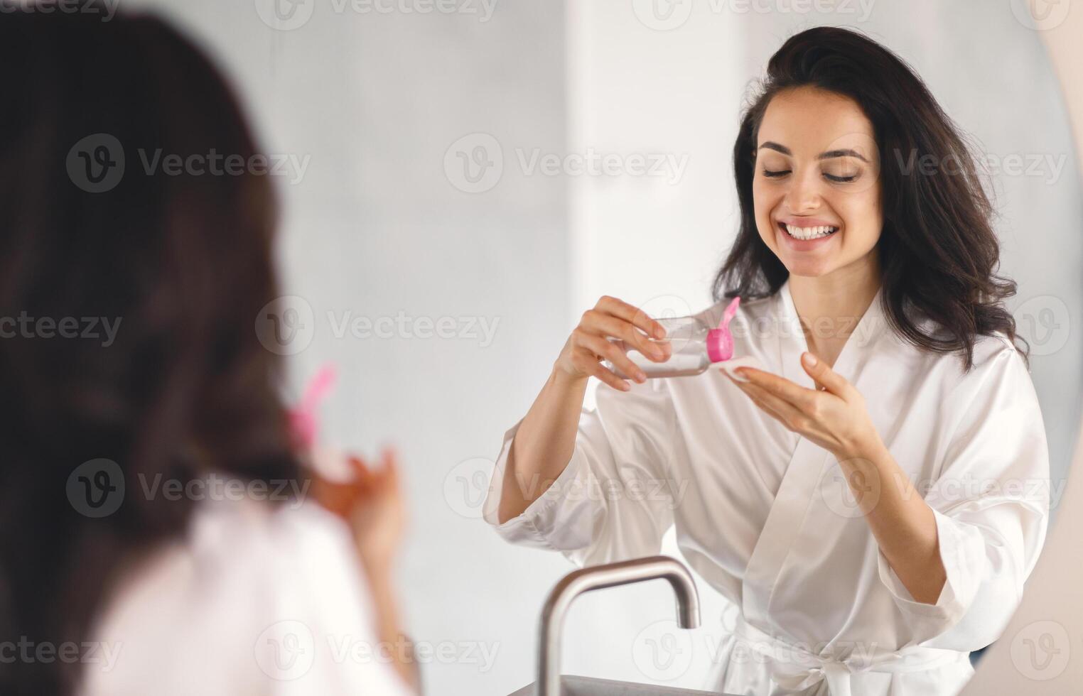 This young Caucasian woman pampering herself in the bathroom shows joy while applying skin care products. She is in a relaxed setting, reflecting self-care and beauty routines. photo