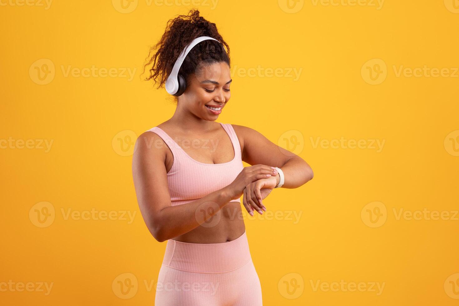A smiling woman in pink athletic clothing adjusts her smartwatch and enjoys music through headphones. The bright yellow background enhances her energetic vibe in a fitness setting. photo