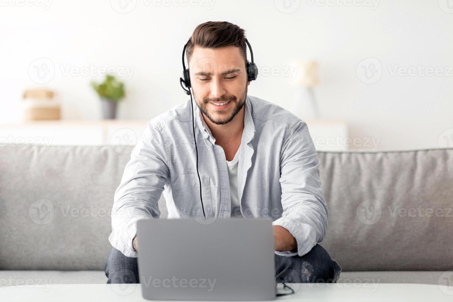 Distance learning concept. Positive man in headset studying online via laptop, sitting on sofa at home, copy space. Handsome guy sitting on couch and watching webinar, enjoying online education photo