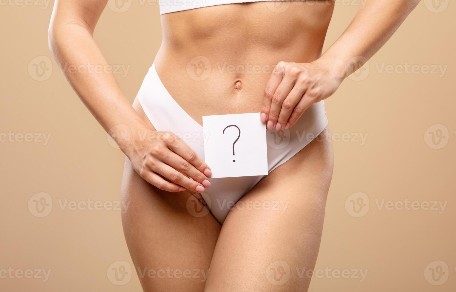 A young Caucasian woman poses confidently in white lingerie, holding a card with a question mark in front of her midsection. The background is a warm, solid color that enhances the focus on her. photo