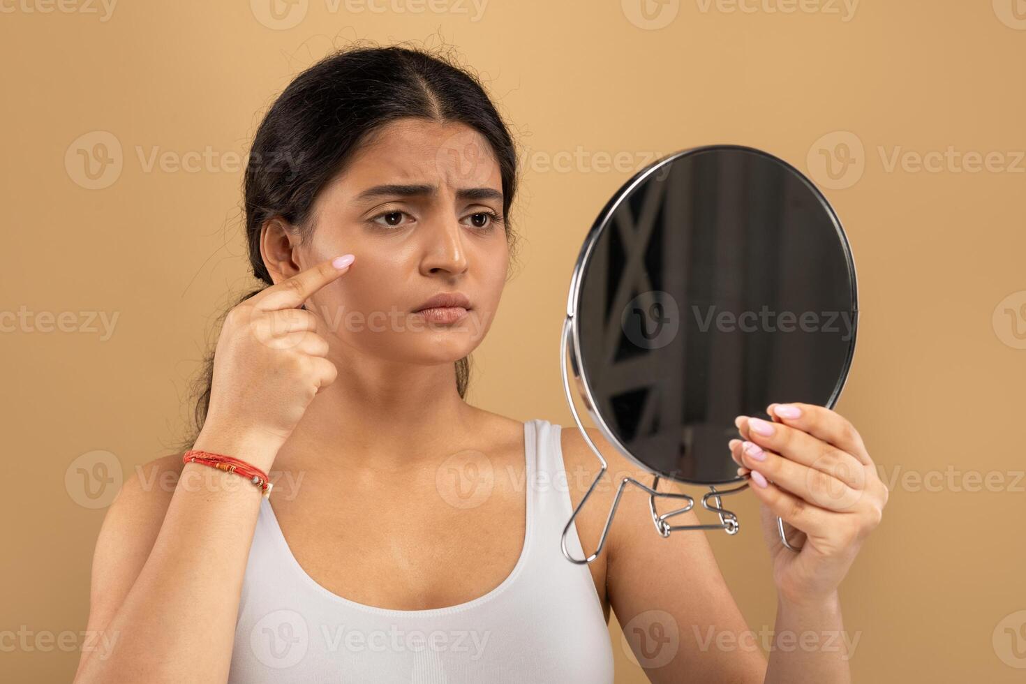In a warm-toned indoor space, a young Indian woman focuses on her skincare routine, using a mirror to assess her skin. She is thoughtfully engaged in self-care activities. photo