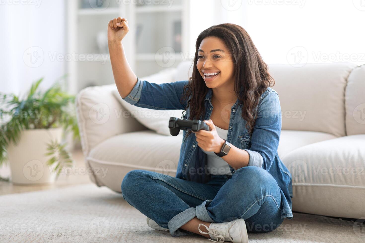 A joyful woman sits on a couch, raising her fist in triumph while playing a game. She is dressed casually, enjoying her time in a comfortable living room setting during the weekend. photo