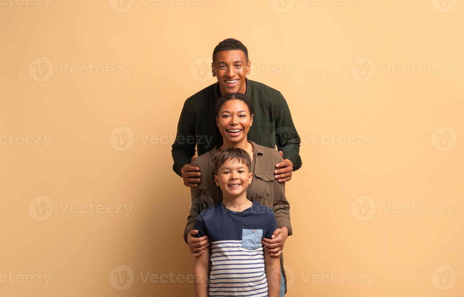 A joyful African American family stands together in a close embrace. A smiling father and mother flank their cheerful teenage son, all exuding love and warmth against a neutral background. photo