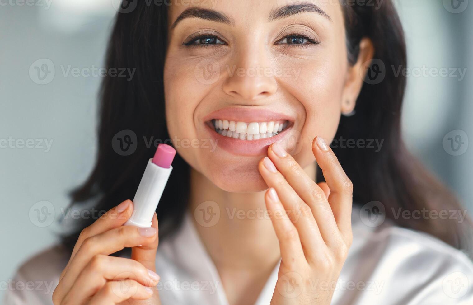 A young brunette woman smiles while applying lip balm in her stylish bathroom. She enjoys a moment of self-care, enhancing her beauty routine with a cheerful expression. photo