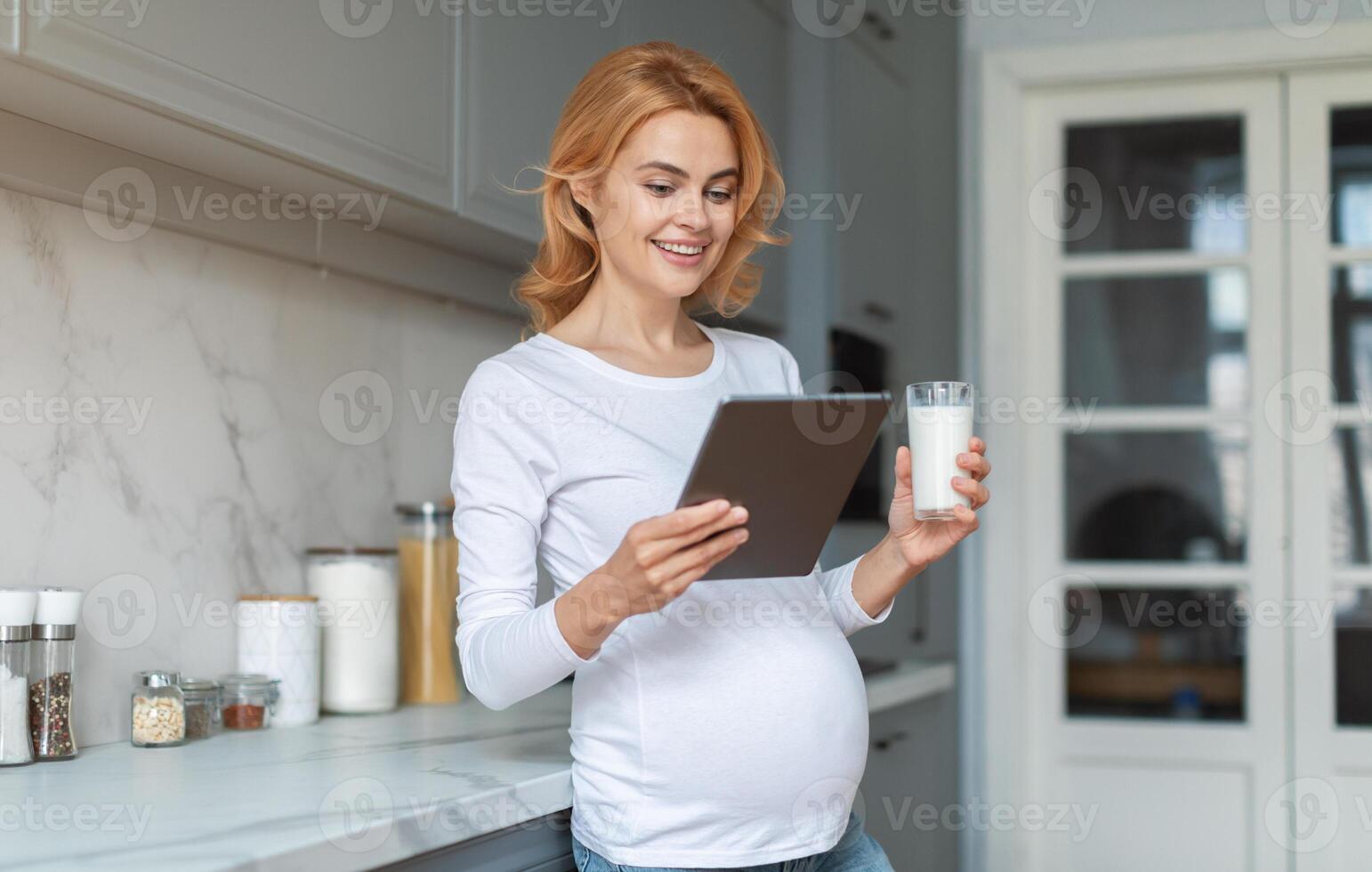 A pregnant woman stands in a bright kitchen, smiling as she looks at a tablet in her hands while holding a glass of milk. The cozy setting features modern decor and natural light. photo