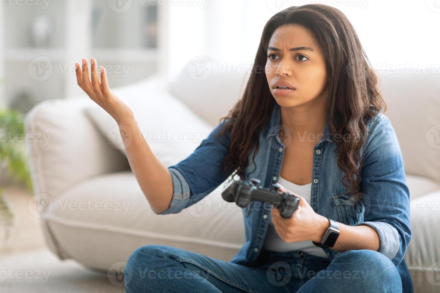 A young black woman sits on a comfortable couch in a bright living room, holding a gaming controller with a confused expression. It appears she is experiencing a challenging moment in her game. photo
