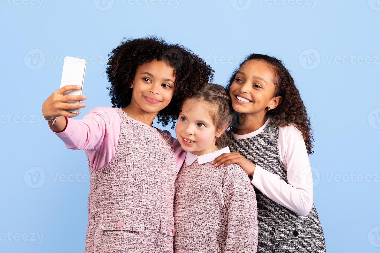 Three happy preteen girls pose closely together, smiling as they take a selfie against a light blue background. Their joyful expressions capture the essence of childhood and friendship. photo
