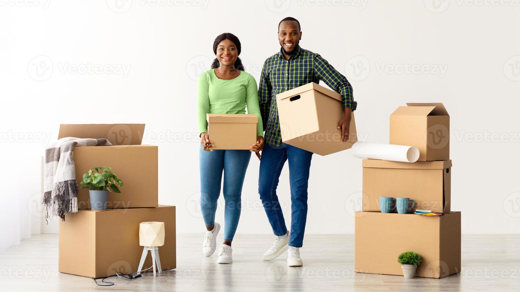 Relocation Concept. Portrait Of Cheerful Black Spouses Moving To New Apartment, Happy African American Couple Holding Belongings, Standing Among Cardboard Boxes And Smiling At Camera, Free Space photo