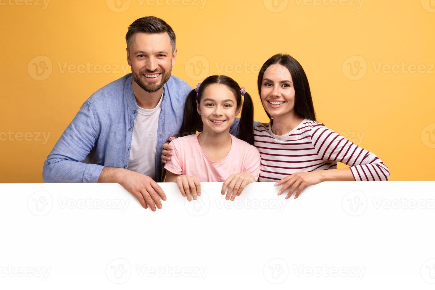 A mother, father, and their teenage daughter smile brightly while leaning on a blank white surface. The cheerful setting emphasizes their connection and warmth, showcasing a loving family bond. photo