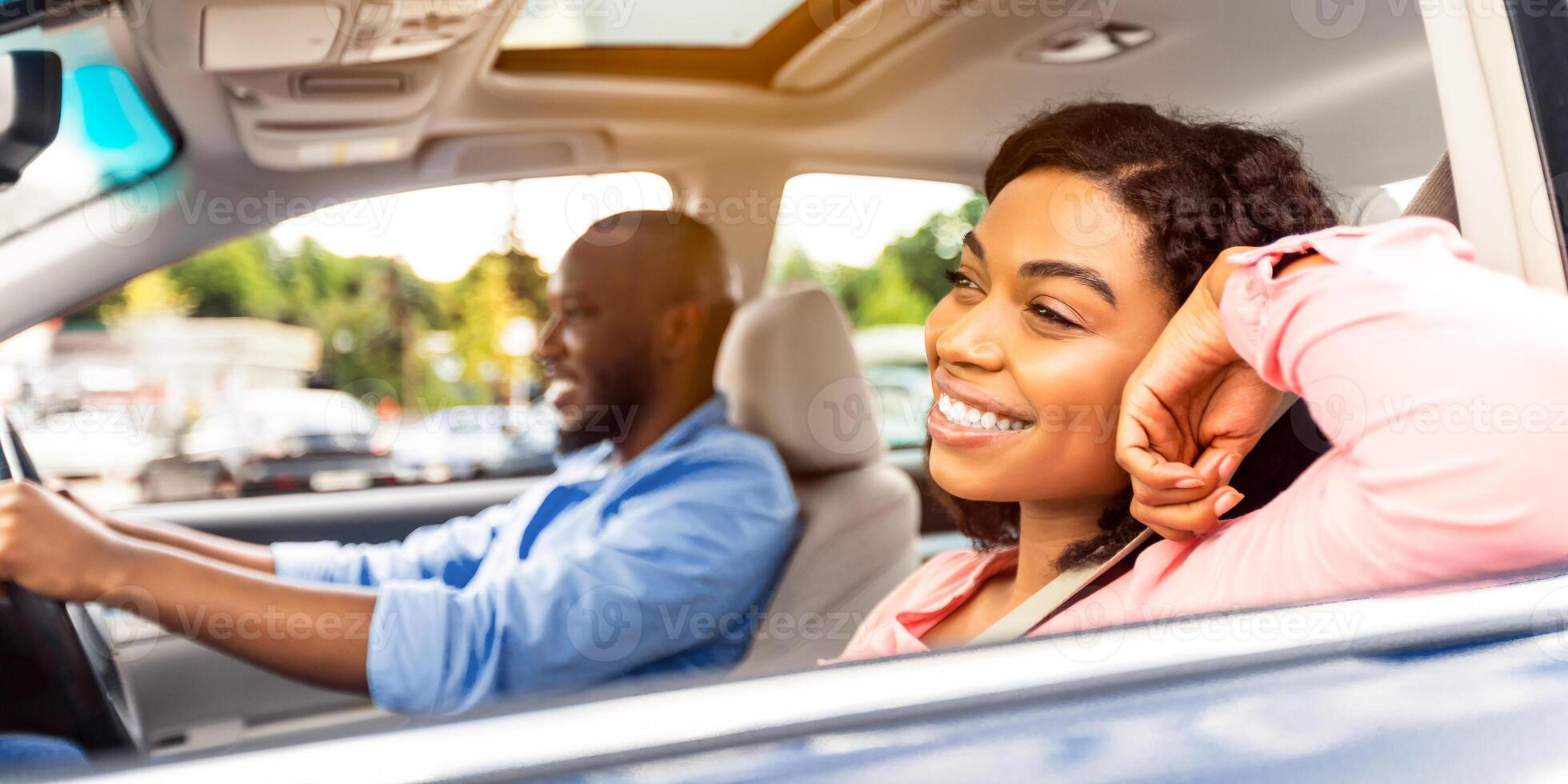 Time For Vacation. Portrait of relaxed cheerful African American woman sitting on the front passenger seat and looking out open window, man driving modern luxury car, enjoying road trip photo