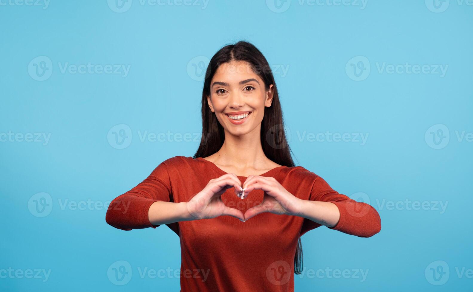 A joyful millennial woman with dark hair forms a heart shape with her hands, smiling brightly against a solid blue backdrop. Her expression radiates happiness and warmth. photo