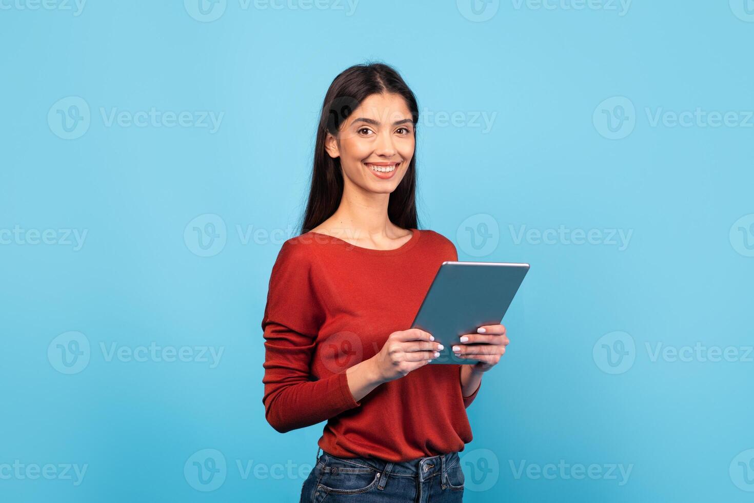 A millennial woman with long brunette hair wears a red sweater and smiles while holding a tablet. The bright blue background enhances her cheerful demeanor, creating a lively atmosphere. photo
