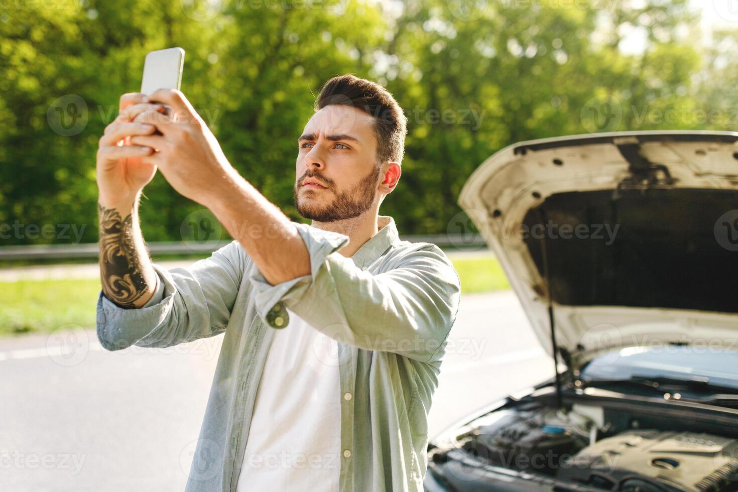 No connection, car breakdown, bad mobile network concept. Sad millennial man standing on road next to broken auto, raising hand with cellphone, trying to call roadside assistance without success photo