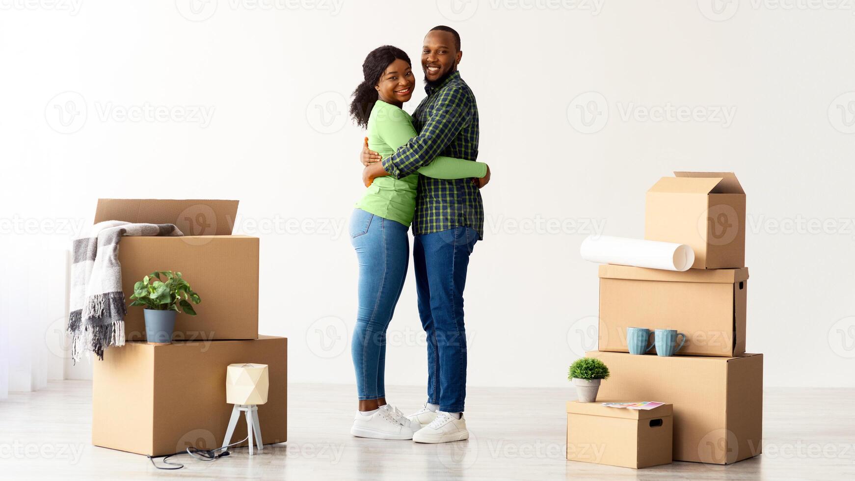 Happy Young Black Spouses Embracing At New Home Among Cardboard Boxes With Belongings After Moving, Portrait Of Cheerful Loving African American Couple Celebrating Relocation, Copy Space photo