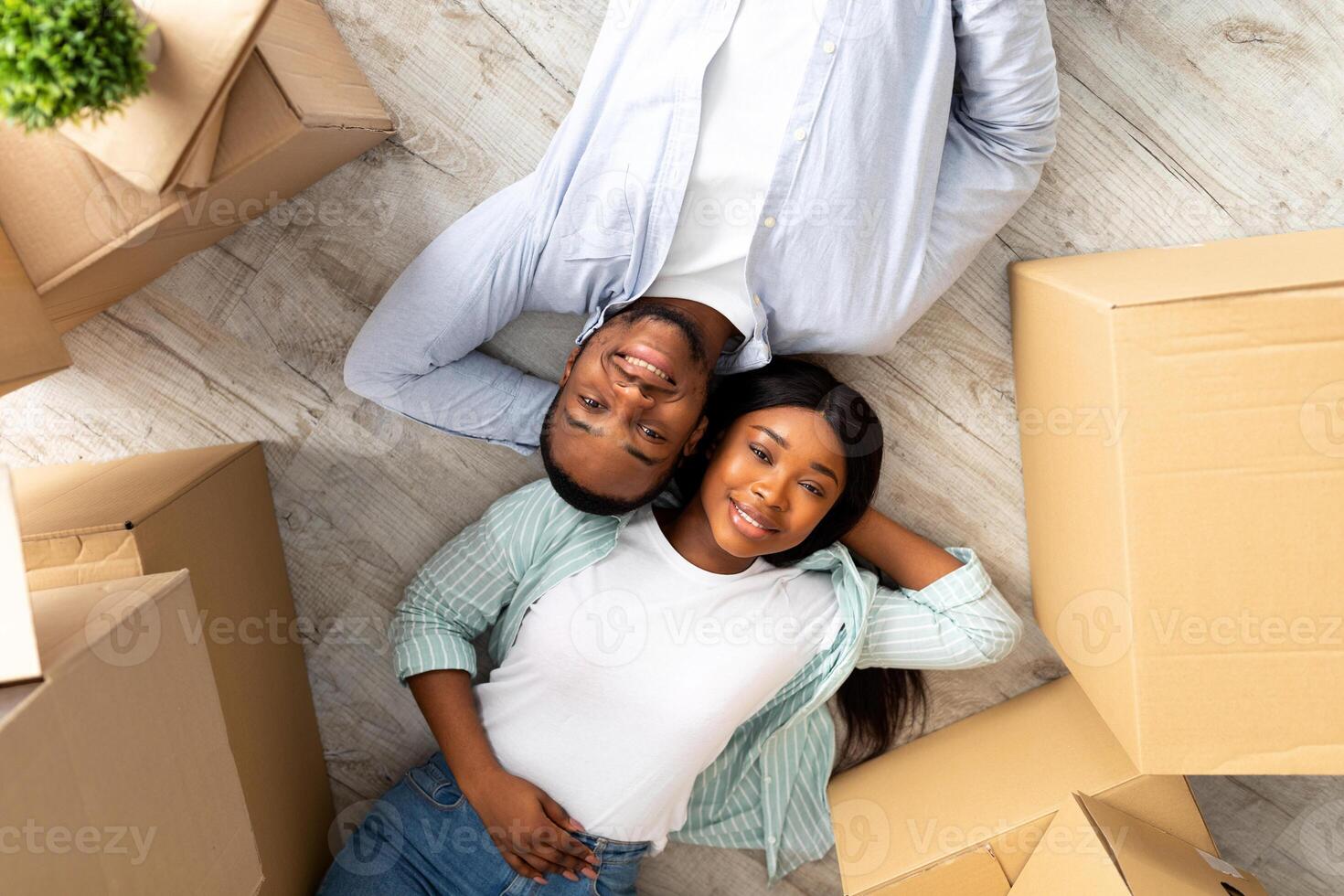Loving african american couple lying among cardboard boxes on floor of their new apartment, top view. Happy black spouses taking break on relocation day and smiling to camera photo