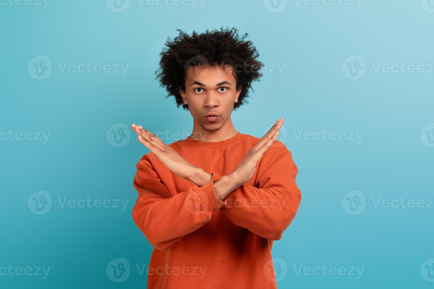 A young African American man with curly hair stands against a light blue background, showcasing a no gesture with crossed arms. The setting emphasizes a vibrant lifestyle and personal expression. photo