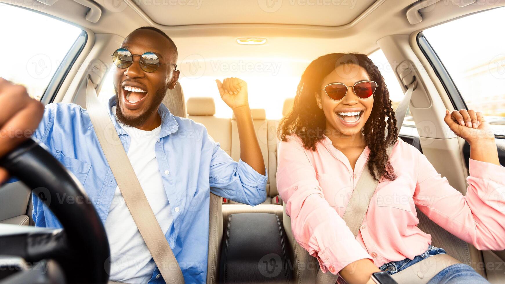 Summer Holidays And Roadtrip Concept. Front view portrait of excited black couple in sunglasses driving car, dancing to music and singing favorite song, going on vacation for rest and relax, sun flare photo