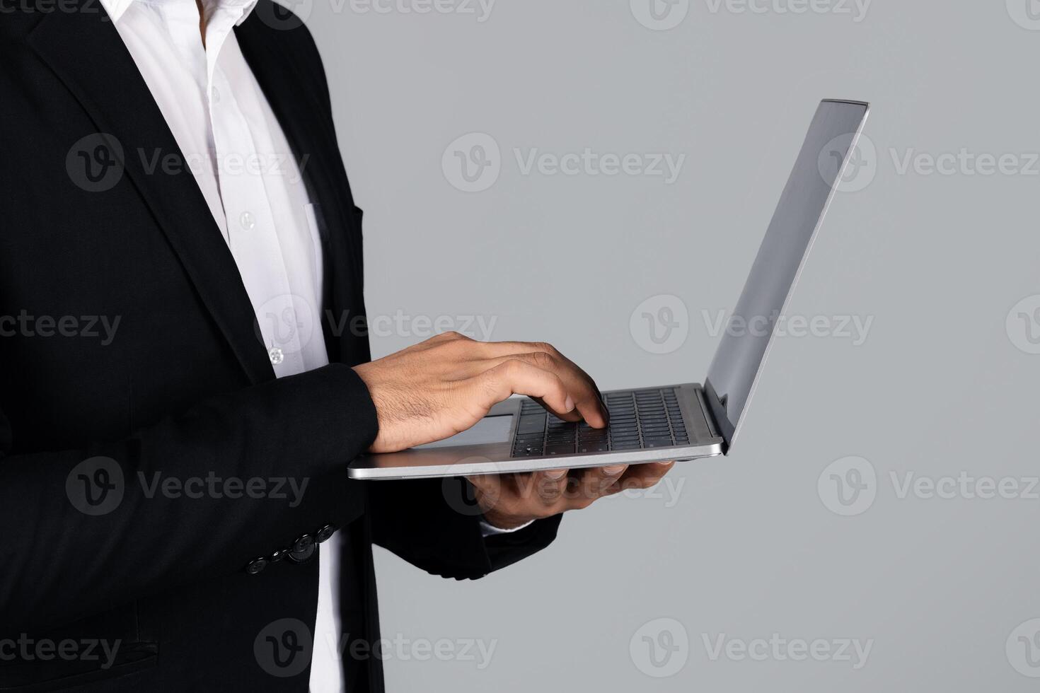 Cropped of entrepreneur stands in a studio environment, focused on typing on a laptop. Dressed in formal attire, he represents a modern, corporate lifestyle. photo