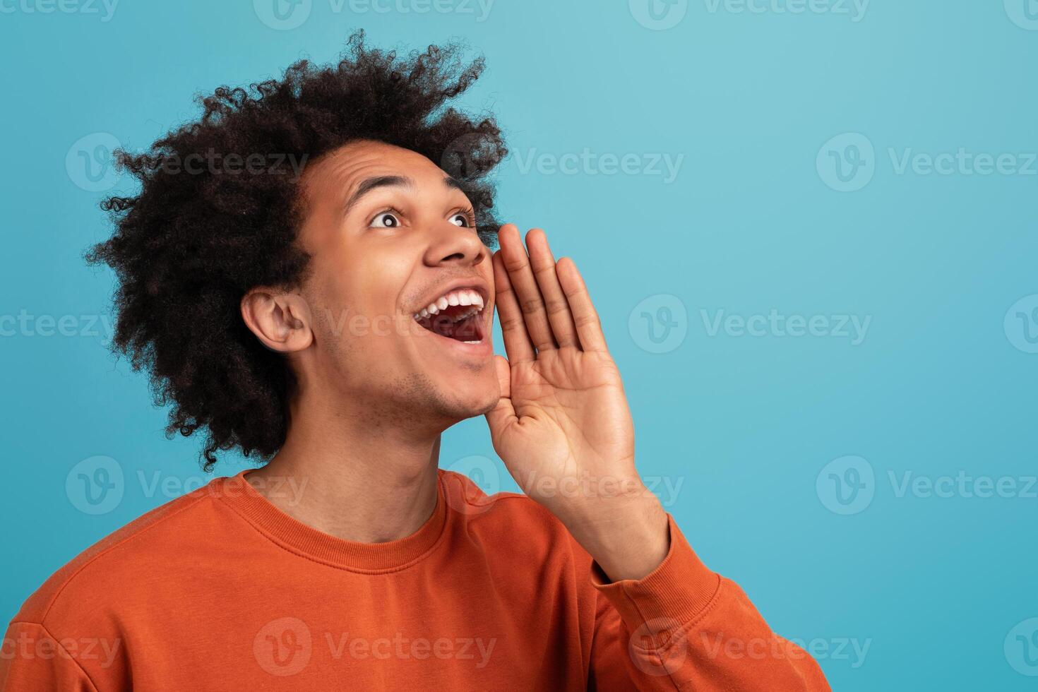 A young African American man with curly hair is joyfully shouting, showing enthusiasm. He stands isolated against a vibrant blue background, capturing a lively moment of expression, copy space photo