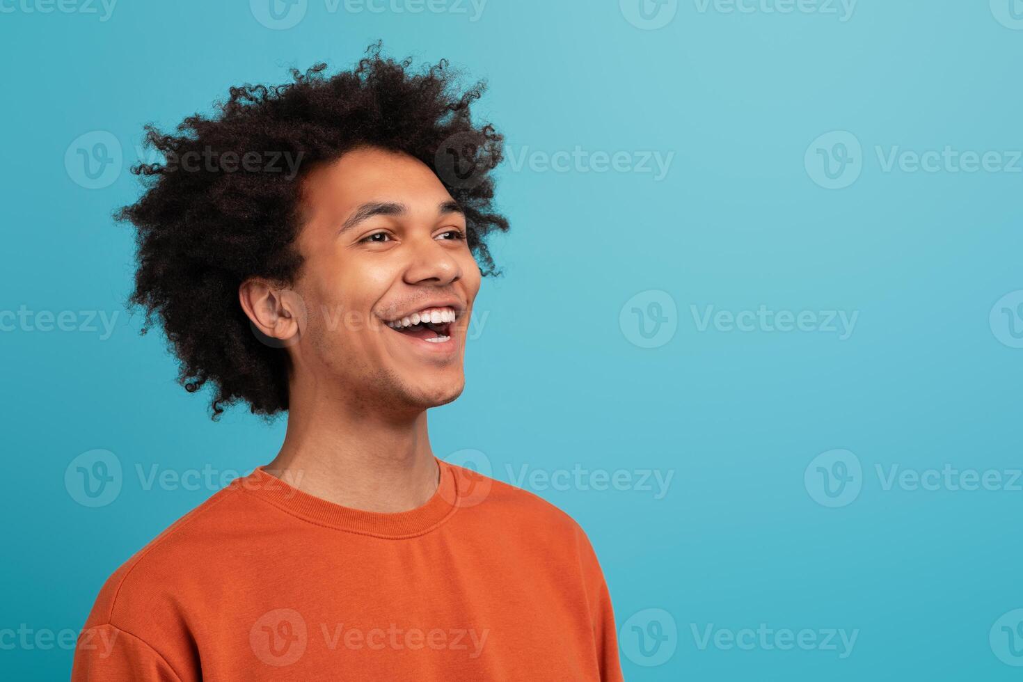A cheerful young African American man with curly hair laughs joyfully. He is dressed in a casual orange sweater and is standing in front of a vibrant blue background, copy space photo