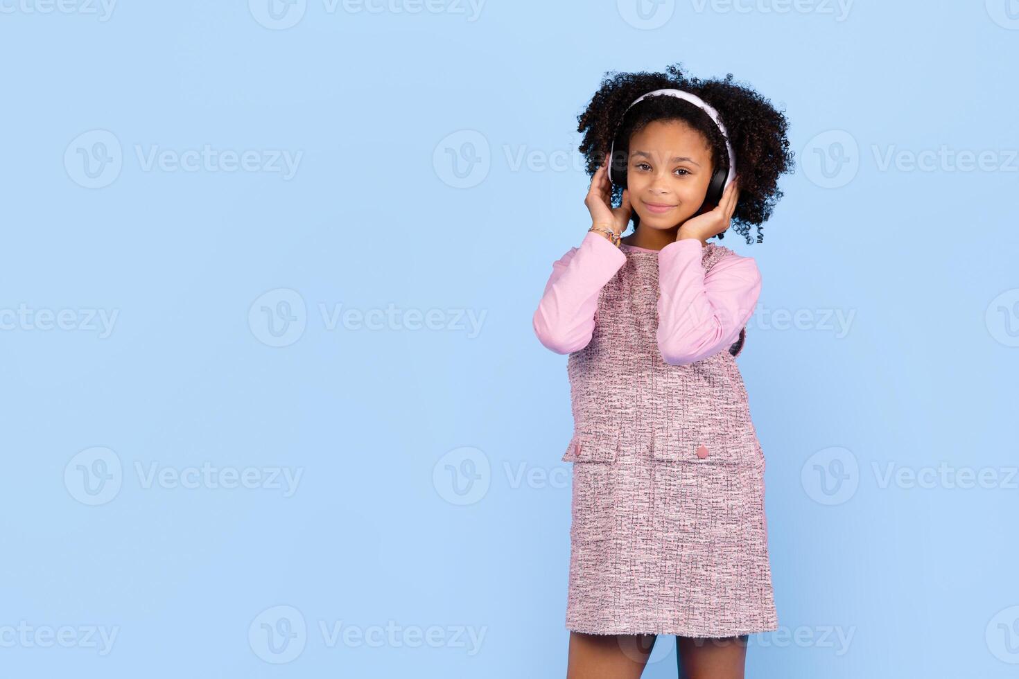A preteen black girl with curly hair smiles while wearing headphones. She is dressed in a stylish outfit and stands in front of a vibrant blue background, radiating joy and excitement, copy space photo