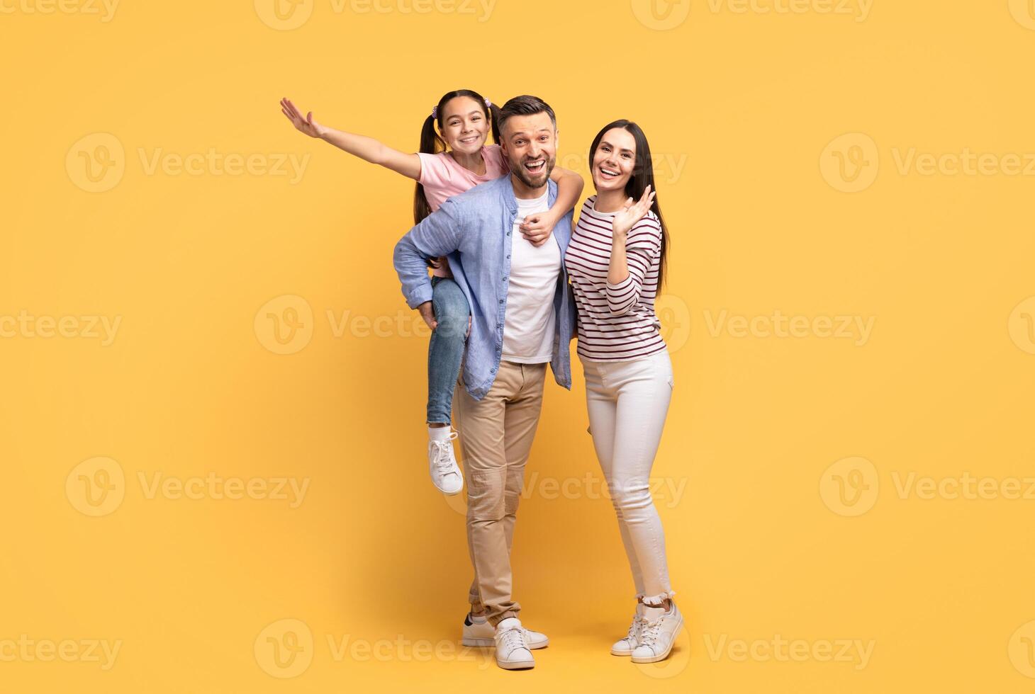 A joyful family poses together against a yellow backdrop. The father carries their daughter on his back while the mother smiles proudly. Love and togetherness are evident. photo
