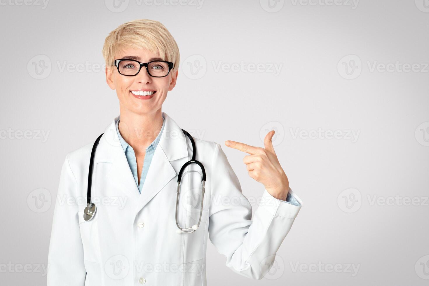 Medical professionals and prevention of disease. Smiling middle aged woman doctor in glasses in white coat with stethoscope, points at herself with finger, isolated on gray background, free space photo