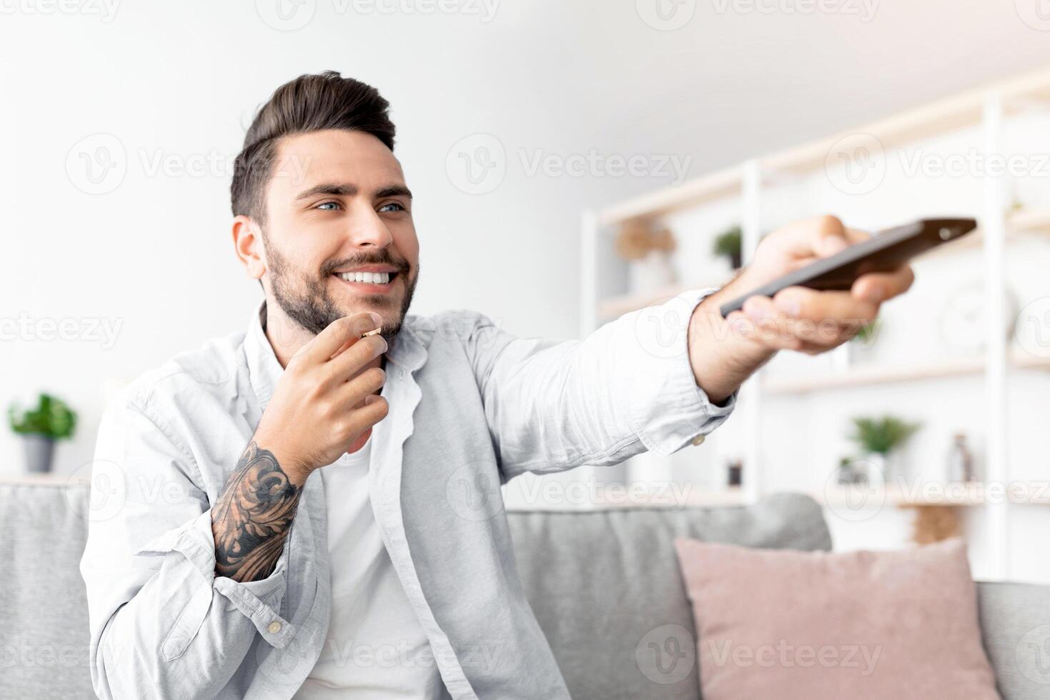 Cheerful millennial guy watching TV and eating popcorn, switching channels with remote controller while relaxing on sofa in living room interior, copy space. Free time concept photo