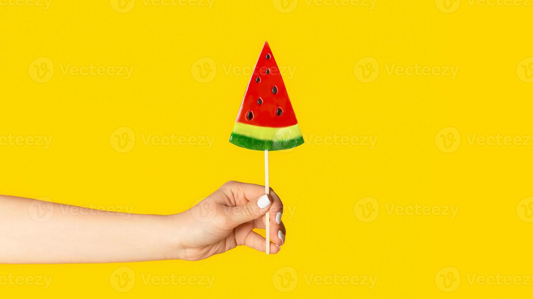 Millennial woman holding watermelon shaped lollipop on orange background, closeup of hand. Copy space. Panorama photo