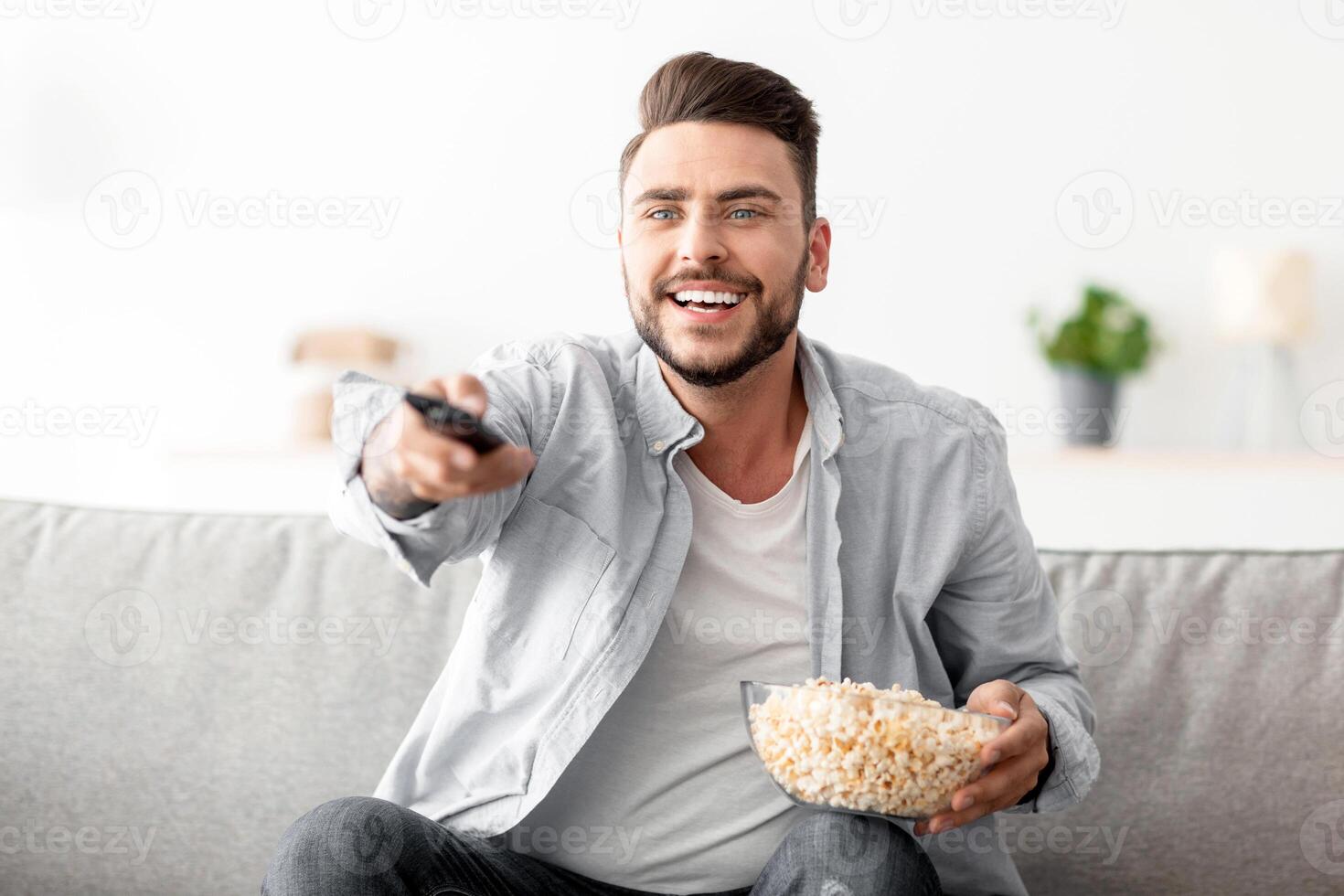 Domestic pastime. Cheerful man resting on couch switching channels with remote controller and eating popcorn, searching his favorite television show, having domestic leisure, copy space photo