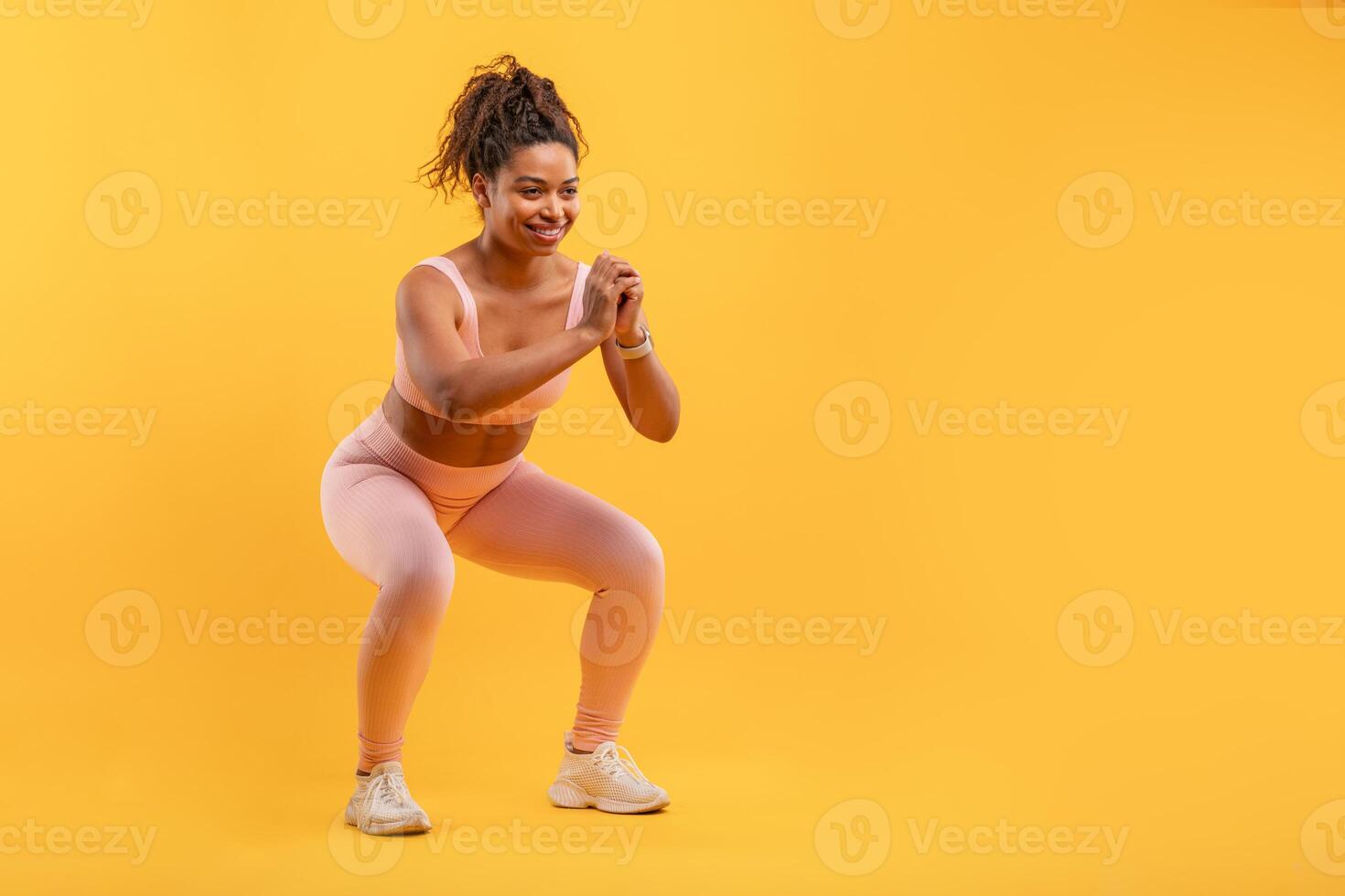 A woman wearing light pink workout attire is energetically doing squats in a vibrant yellow studio. She focuses on her form while exercising with enthusiasm and determination, copy space photo