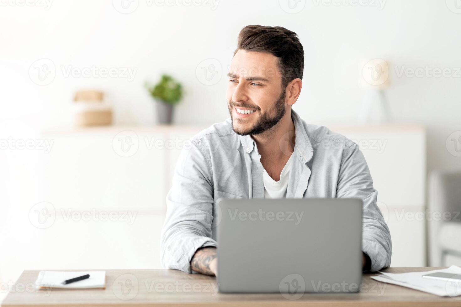 Happy freelance man working on laptop in home office, sitting on workplace and looking aside at free space. Smiling guy thinking about successful business strategy or dreaming about holiday photo