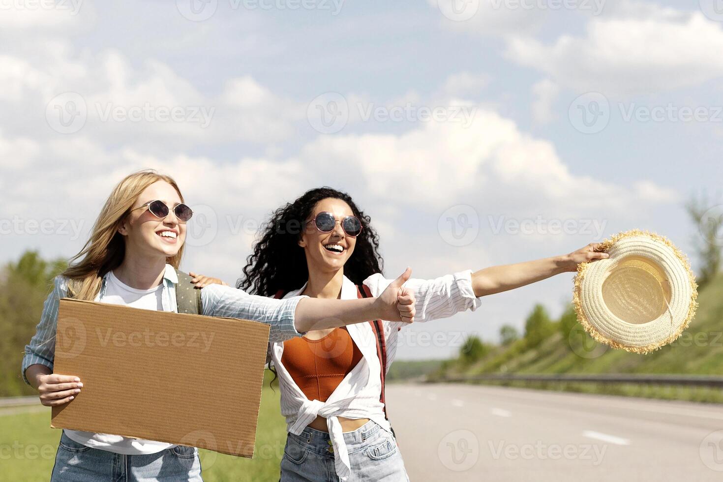 Happy multiracial friends hitchhiking, trying to stop car with empty carton sign, mockup. Group of millennial people with guitar waving down auto on highway. Traveling on summer vacation photo