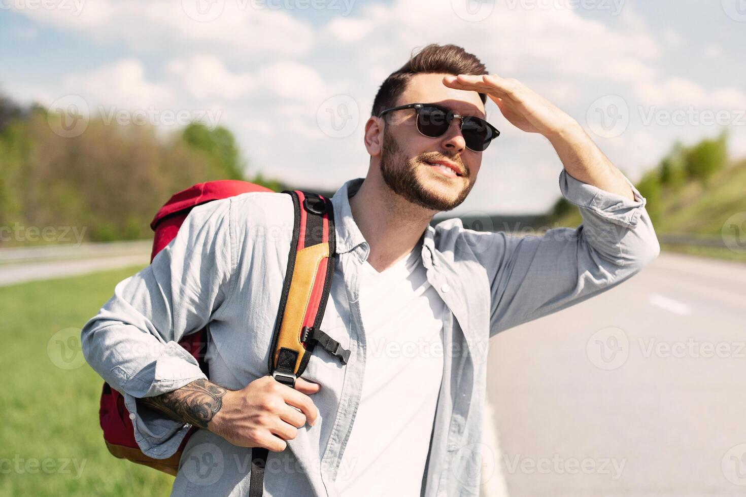 Handsome young man with backpack hitchhiking on road, shielding eyes from sun, looking into distance at passing car, copy space. Autostop journey, summertime travel concept photo