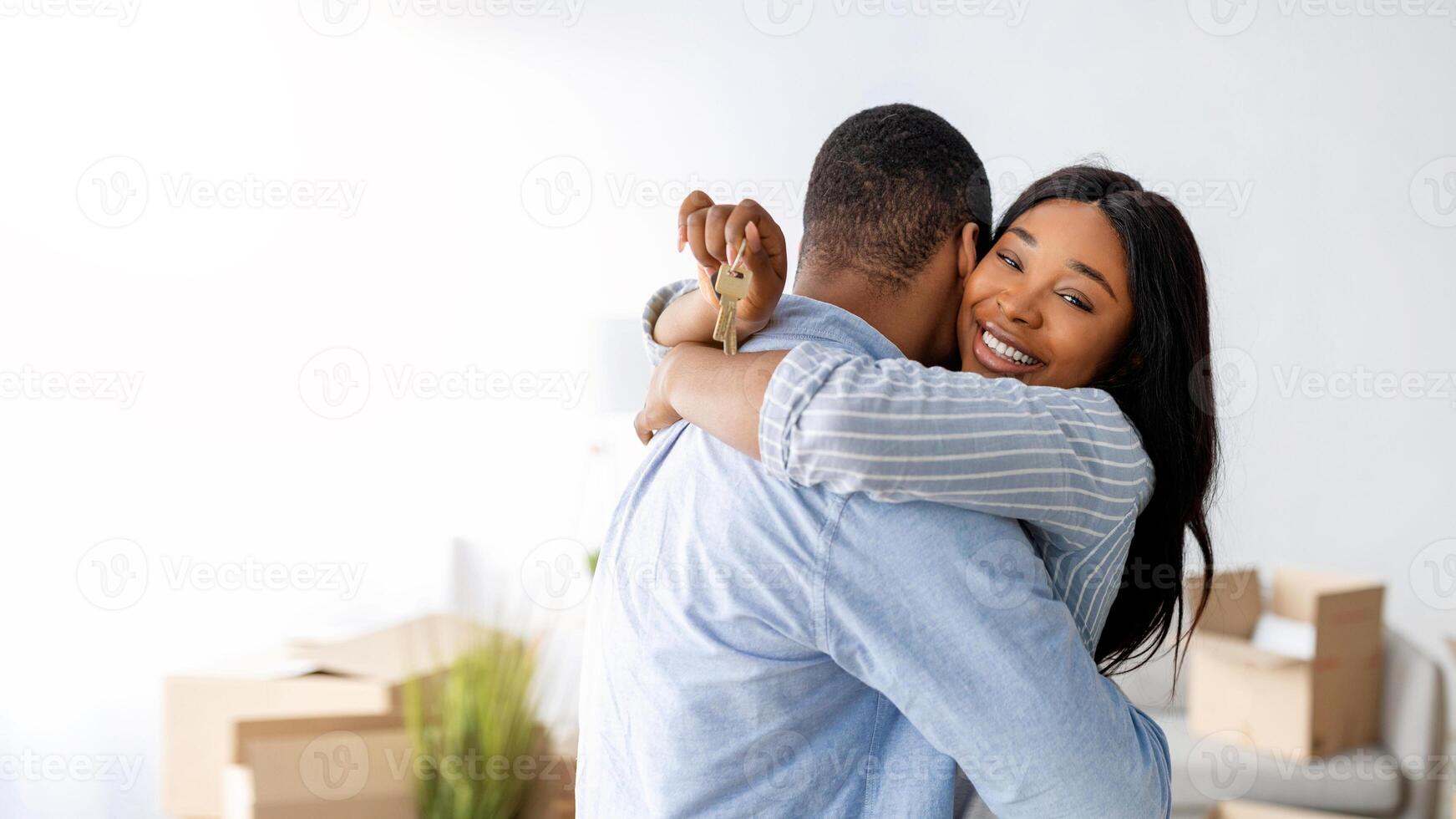 Excited black woman holding house key and embracing her husband, standing in their apartment, free space. Young spouses happy over moving to new property photo