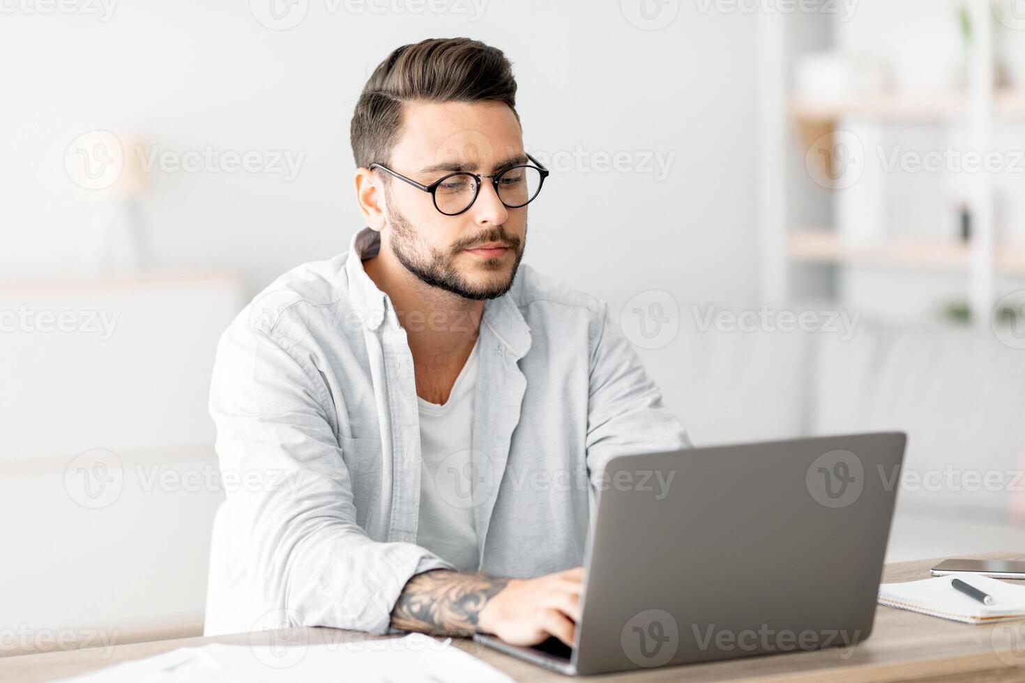 Concentrated self-entrepreneur working with laptop at home office, sitting at desk and using pc computer for remote work, copy space. Male freelancer enjoying distant job opportunities photo