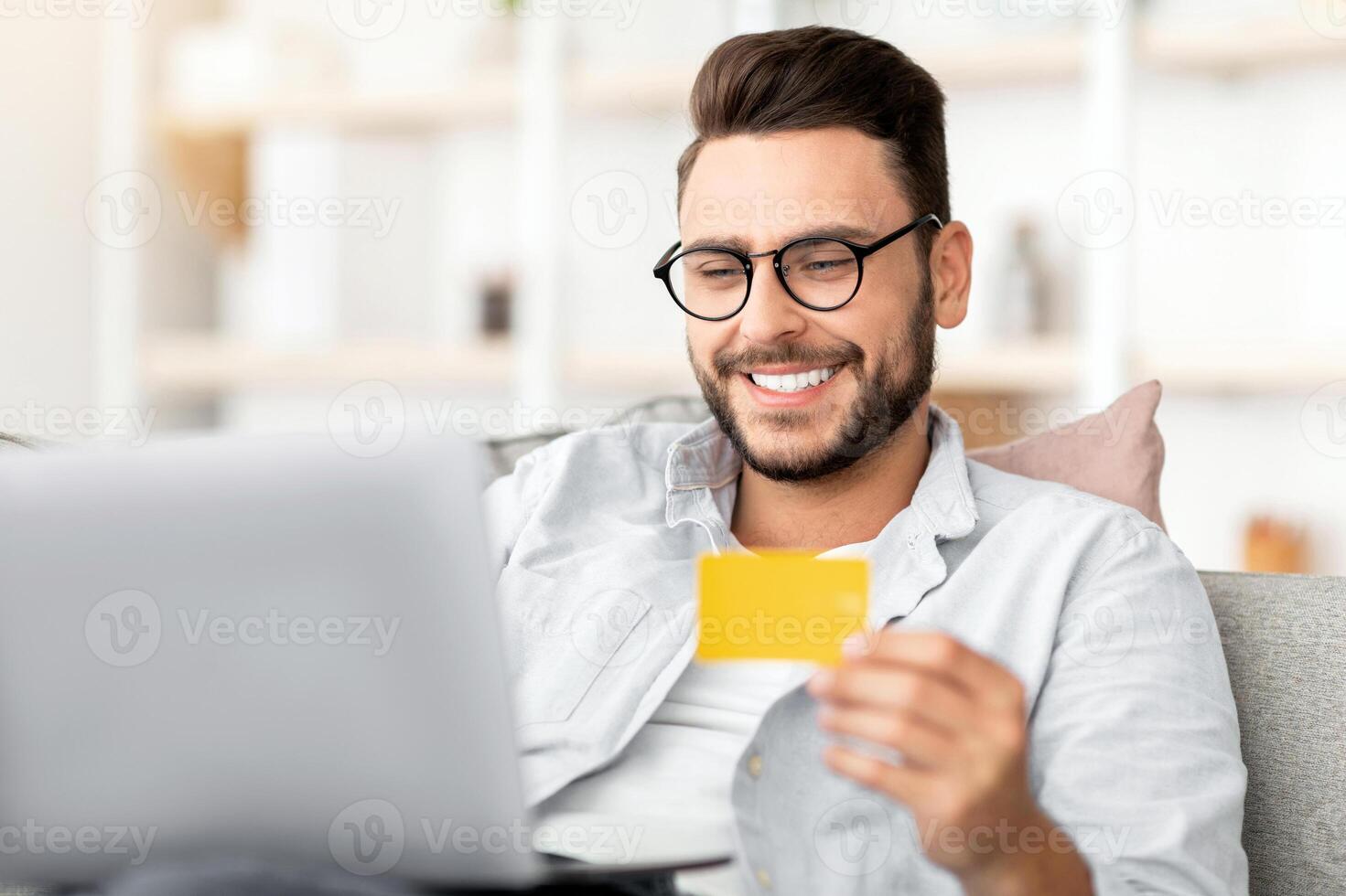 Easy payment and cashback concept. Smiling young man holding credit card and using laptop computer, sitting on couch at home and resting on weekend, shopping online on pc photo