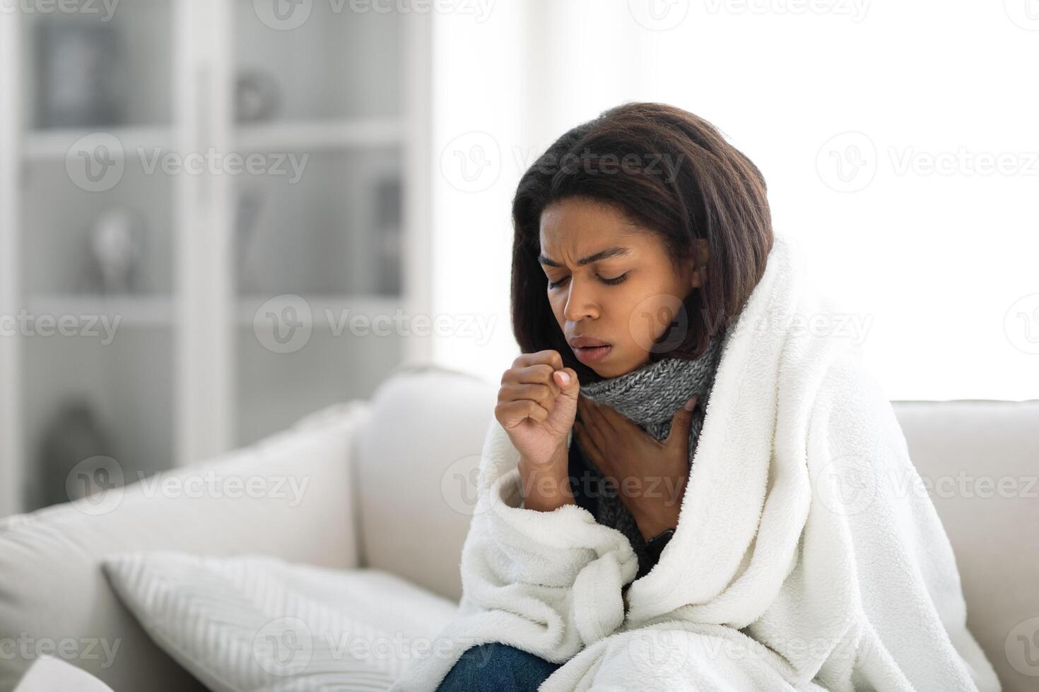A woman is sitting on a sofa, wrapped in a blanket, looking unwell. She is coughing and appears to be resting in her bright, cozy living room during a chilly day, copy space photo
