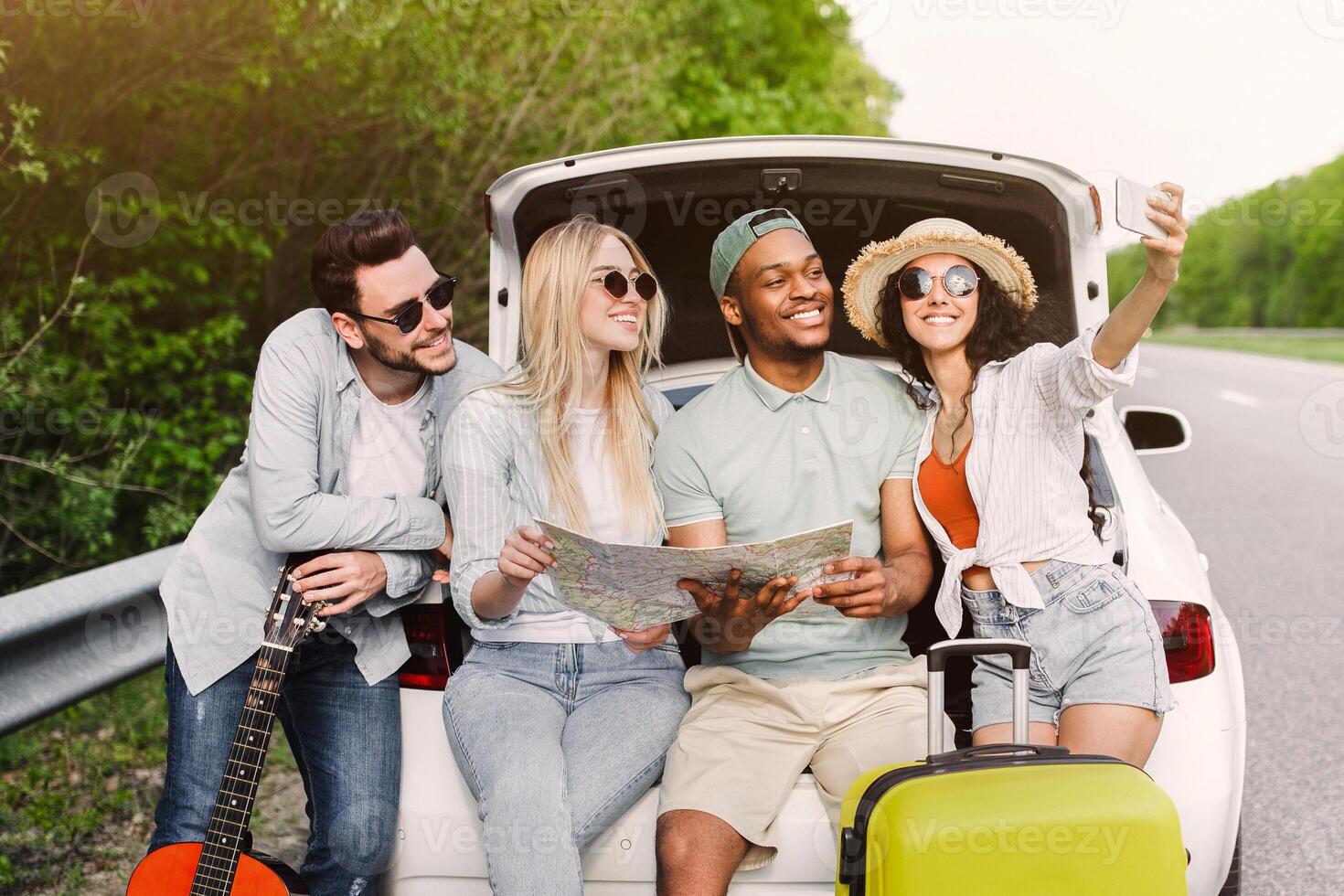 Group of best friends checking map during road trip, sitting in car trunk, taking selfie on their journey. Multiracial young people resting after long drive and having fun. Hitchhiking concept photo
