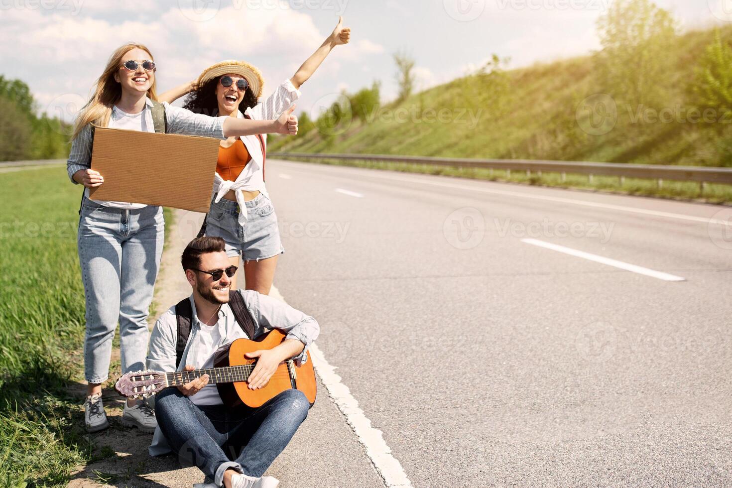 Group of young friends thumbing their ride on road with empty sign, playing guitar, having autostop journey, free space. Millennial hitchhikers having fun road trip, enjoying summer adventure photo