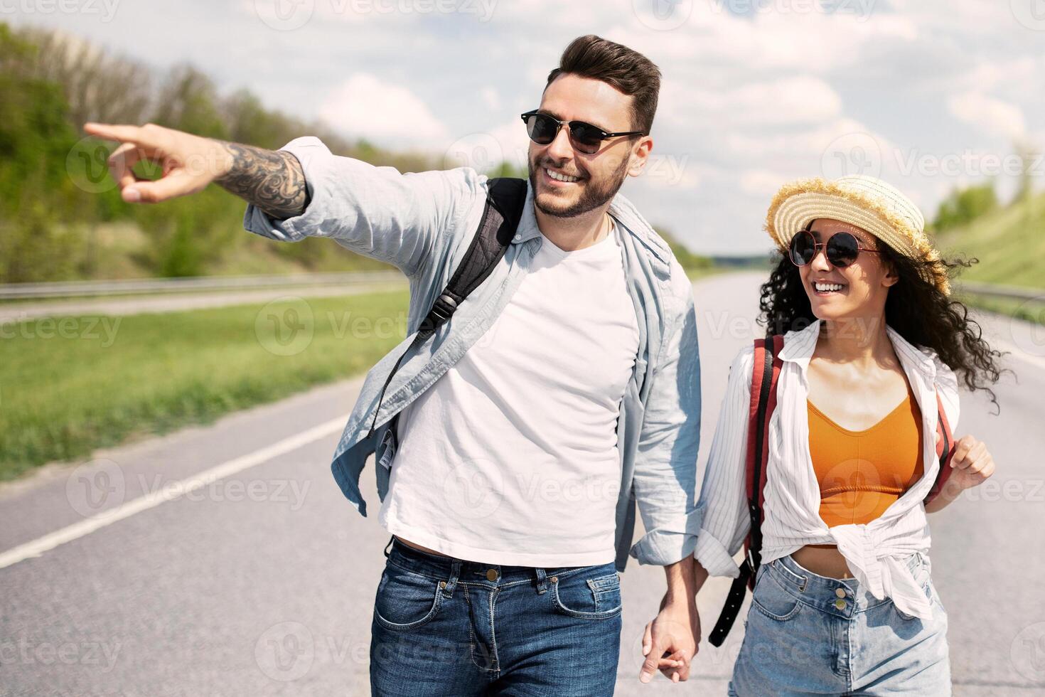 Cheerful young couple with rucksacks walking along road, pointing aside, holding hands, hitchhiking together in summer. Happy guy and his girnfriend enjoying sunny day, having autostop journey photo
