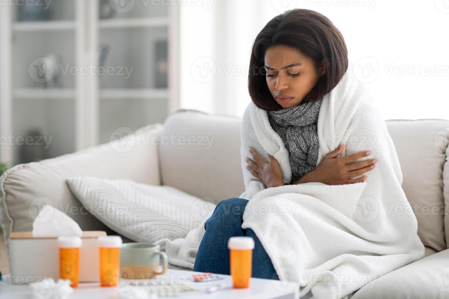 A woman sits on a couch wrapped in a blanket, looking cold and unwell. Medication bottles and tissues are on the table nearby, highlighting her struggle with illness during a cold day, copy space photo