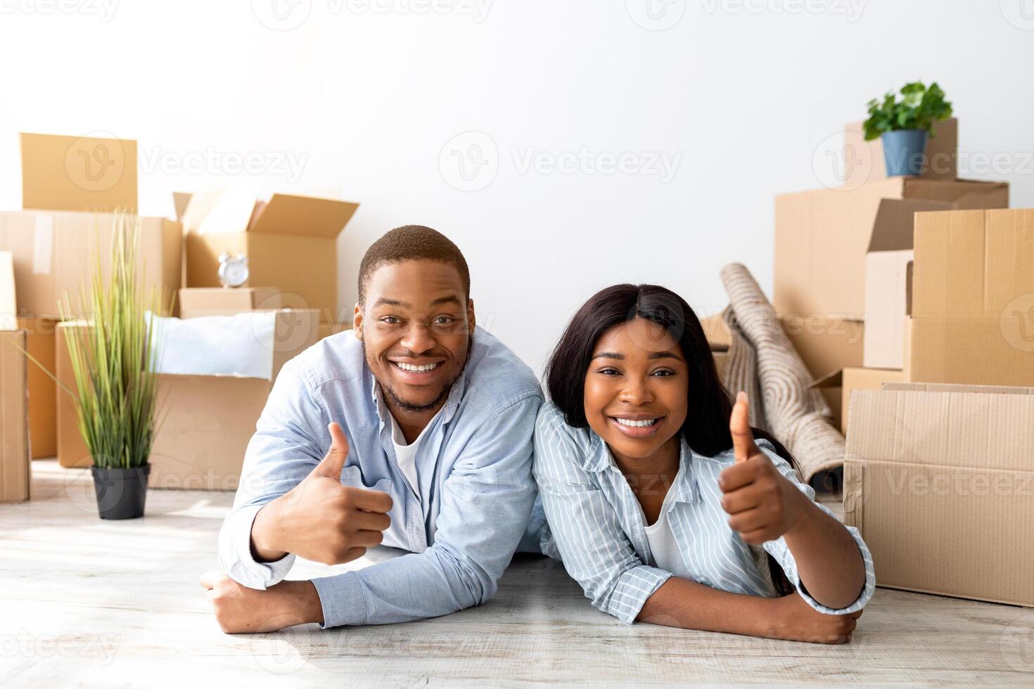 Happy black couple recommending real estate agency, showing thumbs up while lying on floor in new apartment among cardboard boxes and smiling to camera photo