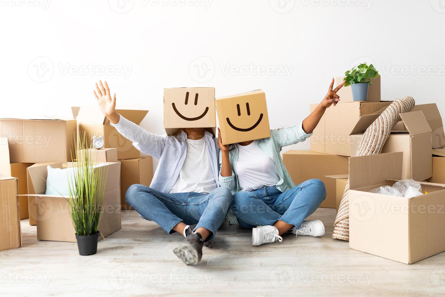 Joyful black couple with cardboard boxes on heads, sitting on floor and having fun, celebrating moving day, relocation to new apartment. Home, people, moving and real estate concept photo