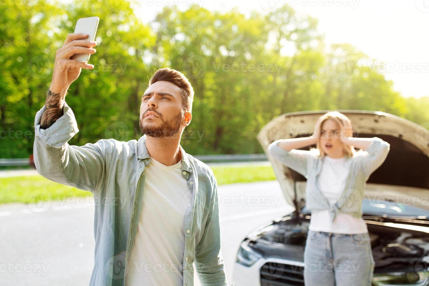 Sad young man standing on road next to his broken car together with terrified girlfriend, raising hand with mobile phone, looking for network signal, trying to call roadside assistance. No connection photo