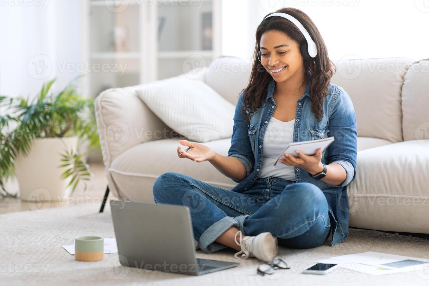 A young black woman, wearing headphones, is actively participating in an online class while seated on a couch. She appears focused and happy, holding a tablet and interacting with her laptop. photo