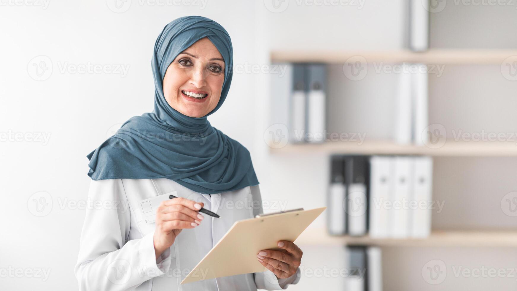 Happy Muslim Female Doctor Posing Smiling To Camera Holding Medical Case Record Standing In Modern Clinic Office Indoors. Portrait Of Mature Middle-Eastern Professional Therapist Lady Wearing Hijab photo